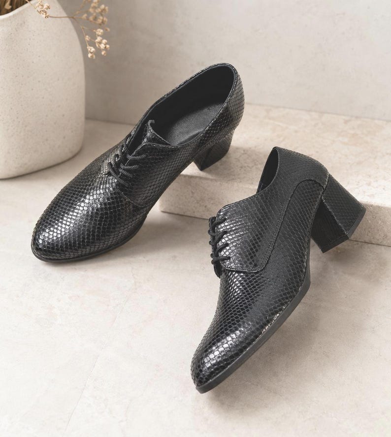 May include: A pair of black, textured leather shoes with a lace-up design and block heels. The shoes have a snakeskin pattern and are displayed on a neutral surface. The heels appear to be approximately 2 inches high.