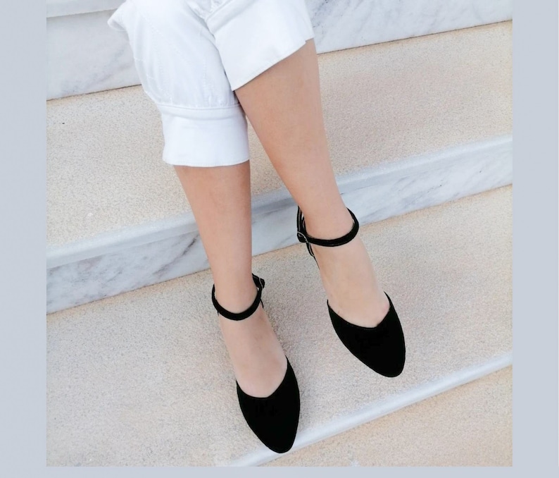 May include: A pair of black suede ankle strap shoes with a pointed toe. The shoes are worn by a person sitting on a set of marble stairs.