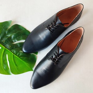 May include: A pair of black leather Oxford shoes with a pointed toe and lace-up closure. The shoes are displayed on a white surface with a large green leaf in the background. The interior of the shoes is a light brown color.
