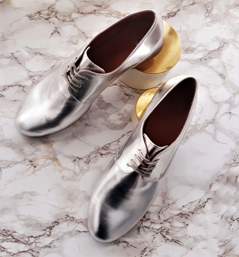 May include: A pair of silver metallic leather oxfords with lace-up closures. The shoes are on a white marble surface.