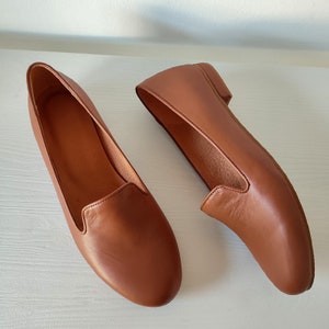 Puede incluir: Un par de mocasines de cuero marr&oacute;n. Los zapatos tienen una punta redondeada y un tac&oacute;n bajo. El cuero tiene una textura suave y un color marr&oacute;n rojizo c&aacute;lido.