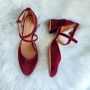 May include: A pair of burgundy velvet heeled shoes with ankle straps. The shoes have a closed toe and a block heel.