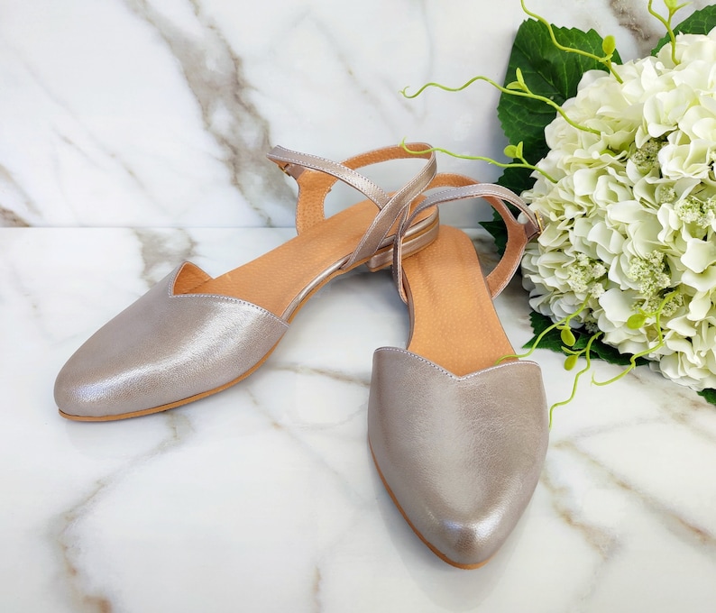 Op de afbeelding: Een paar zilveren metallic leren sandalen met enkelbandjes. De sandalen hebben een spitse neus en een lage hak.