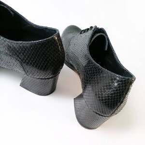 May include: Black leather shoes with a snakeskin pattern. The shoes have a low, block heel and lace-up closure. The heels measure approximately 2 inches.