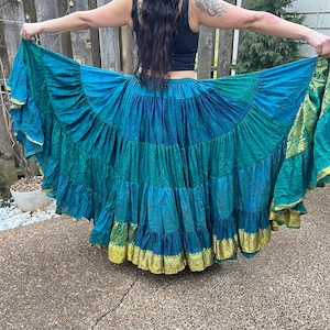 Puede incluir: Una falda fluida de color turquesa y azul con ribete dorado. La falda está hecha de tela en capas con un dobladillo con volantes. El ribete dorado añade un toque de elegancia. La falda se sostiene abierta para mostrar su forma completa.