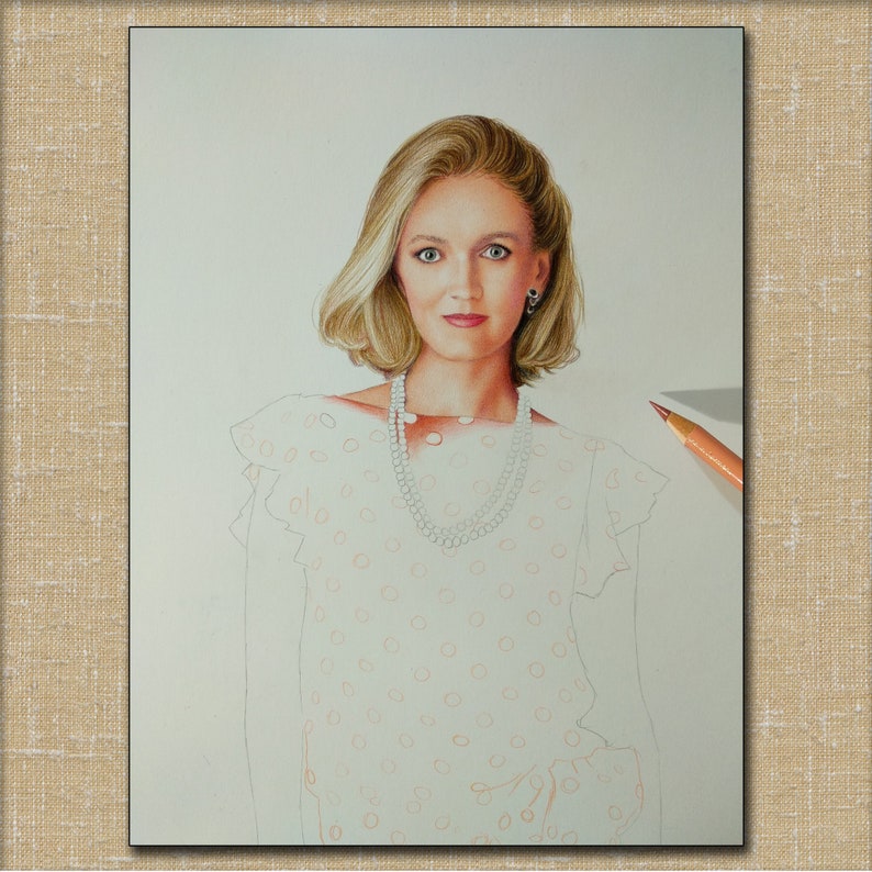 Op de afbeelding: Een vrouw met blond haar draagt een witte blouse met een stippenpatroon. Ze draagt een parelketting en oorbellen. De blouse heeft een gerimpelde kraag en mouwen.