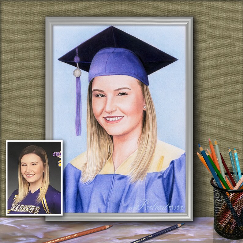 Op de afbeelding: Een kleurpotloodtekening van een persoon die een blauwe afstudeerhoed en -jurk draagt. De persoon heeft blond haar en lacht. De tekening is ingelijst in een zilveren lijst.