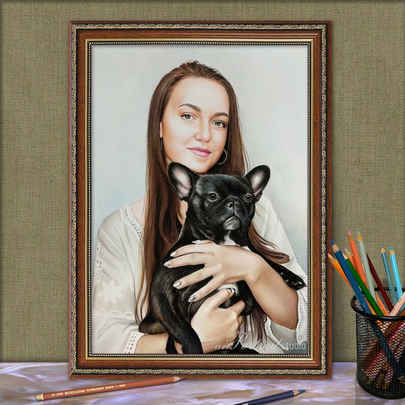Op de afbeelding: Een vrouw met lang bruin haar houdt een zwarte Franse bulldog in haar armen. Ze draagt een wit shirt met een bloemenpatroon. Het schilderij is ingelijst in een bruin houten lijst met gouden accenten.