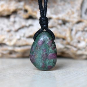 Pode incluir: Um pingente de zoisite polida em forma de gota, com uma superfície manchada em verde e magenta profundo. A pedra está suspensa por um cordão preto simples, preso no topo com um pequeno nó apertado, criando um colar de design funcional.