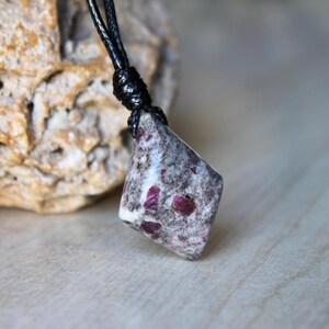 May include: A pendant featuring a polished, diamond-shaped stone with a mottled grey and white base color and embedded deep magenta-red inclusions. The stone is suspended from a black cord that is knotted securely at the top of the pendant.