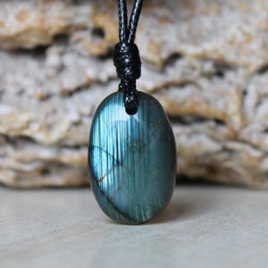 May include: An oval-shaped labradorite pendant featuring a deep blue and teal iridescent sheen with vertical striations. The polished gemstone is suspended from a simple black cord that passes through a drilled hole at the top of the stone.