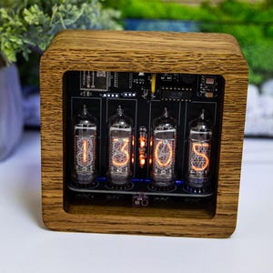 May include: A wooden digital clock displaying the time 1:30:05. The clock has four glass tubes with orange glowing numbers. The clock is encased in a square, light brown wooden frame. The background includes a small bonsai tree and green foliage.