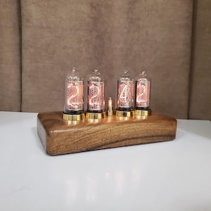 May include: A wooden digital clock with four glass tubes displaying the numbers 2, 4, 2, and 4. The tubes have a warm, orange glow and are mounted on a rectangular wooden base with gold-colored accents. A small light is between the tubes.