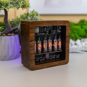 Può includere: Un orologio in legno con un quadrante chiaro che mostra l'ora 2:17:04 usando sei tubi Nixie arancioni incandescenti. L'orologio è in legno scuro e ha un design semplice.