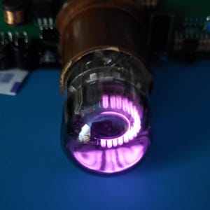 May include: Close-up of a vacuum tube with a glowing purple interior. The glass bulb is clear, revealing internal components. The tube is set against a blue surface, with a circuit board visible in the background. The light emitted is a bright, vibrant purple.