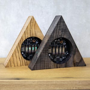 May include: Two triangular wooden clocks with digital displays. One clock is light brown, the other is dark gray. The digital displays show green numbers. The clocks are on a wooden surface.