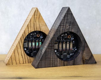 May include: Two triangular wooden clocks with digital displays. One clock is light brown, the other is dark gray. The digital displays show green numbers. The clocks are on a wooden surface.