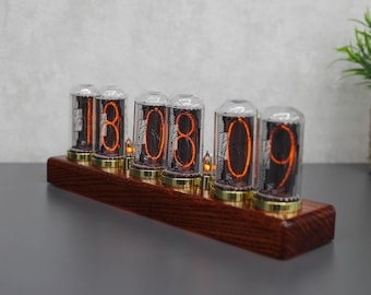 May include: A digital clock displaying "18:08:09". The clock features six clear glass tubes with orange numerals, mounted on a dark wood base with gold-colored accents. Small, decorative lights are placed between the tubes.