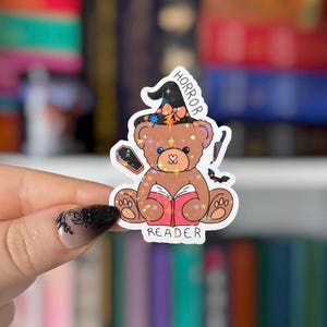 May include: A die-cut sticker featuring a brown teddy bear wearing a black witch hat with an orange bow. The bear holds an open red book and is surrounded by a small coffin, a knife, and a bat. The text reads HORROR READER in black letters.