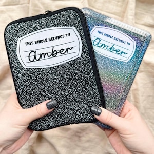 May include: Two black Kindle covers with a white label that says "This Kindle belongs to" and a handwritten name "Amber". One cover is a black and white speckled design and the other is a holographic iridescent design.