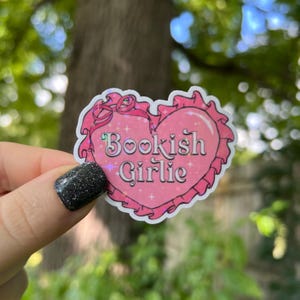 May include: A pink heart-shaped sticker with a glittery finish and the text "Bookish Girlie" in a white font.