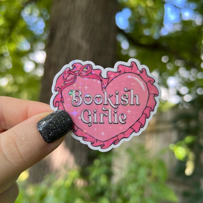 May include: A pink heart-shaped sticker with a glittery finish and the text "Bookish Girlie" in a white font.