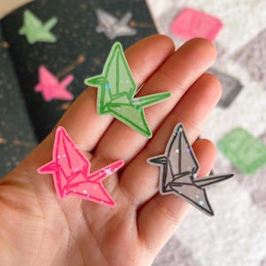 May include: Three origami paper crane stickers with glitter accents. The cranes are pink, green, and grey.