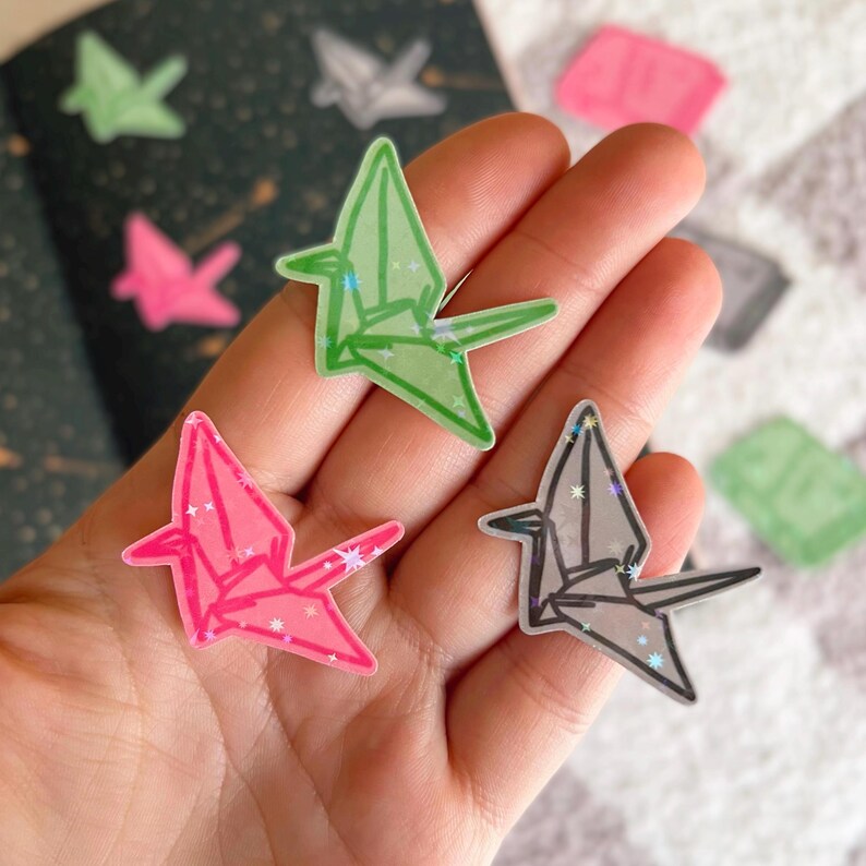 May include: Three origami paper crane stickers with glitter accents. The cranes are pink, green, and grey.