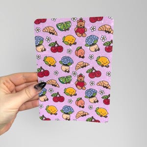 May include: A rectangular sticker featuring a repeating pattern of cartoon fruits, pastries, and a teddy bear on a light purple background. The design includes cherries, lemons, croissants, mushrooms, and flowers. The sticker is held by a hand.