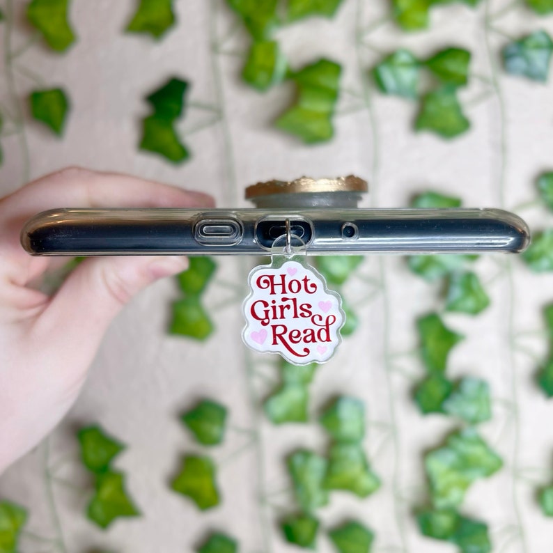 May include: A white phone charm with a pink cloud shape and the text "Hot Girls Read" in red lettering. The charm is attached to a clear phone case.