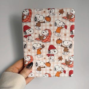May include: A rectangular e-reader case featuring a repeating pattern of Snoopy characters on a light pink and white gingham background. The design includes Snoopy wearing sweaters, scarves, and hats, holding pumpkins, reading books, and autumn leaves.