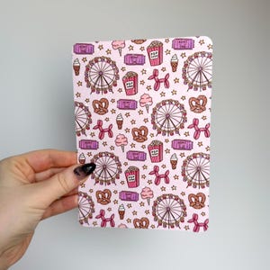 May include: A pink notebook with a repeating pattern of carnival-themed illustrations. The design includes Ferris wheels, popcorn, ice cream cones, pretzels, balloon dogs, stars, and tickets. The notebook is held by a hand.