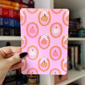 Puede incluir: Una tarjeta rectangular rosa con un patrón repetido de calabazas en marcos ovalados con lazos. Las calabazas son naranjas, blancas y rosas. Los marcos son naranjas y el fondo es rosa liso.
