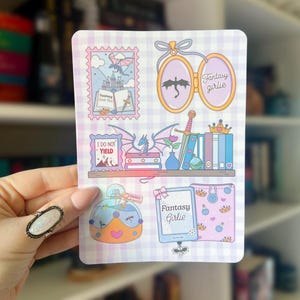 Puede incluir: Una hoja de pegatinas con ilustraciones de temática fantástica. Los diseños incluyen un dragón, libros, una espada y el texto "Fantasy Girlie". La paleta de colores presenta rosas pastel, azules y morados, con detalles dorados. Sostenido por una mano.