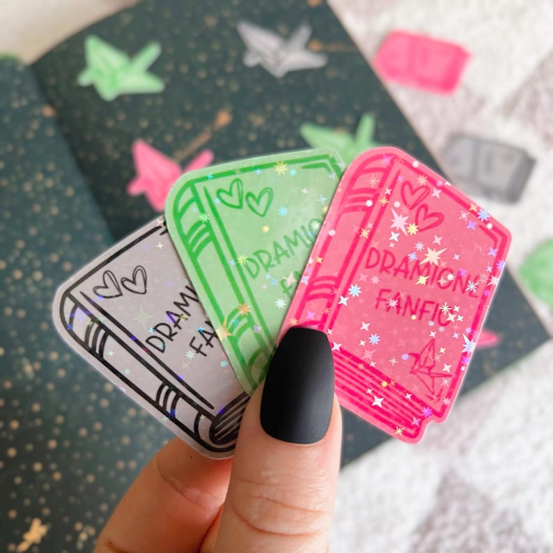 May include: Three holographic book-shaped stickers with glitter and stars. The stickers say "Dramione Fanfic" in different colours.