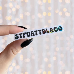 May include: A white sticker with the text "STFUATTBLAGG" in black, bubble letters with rainbow glitter. The sticker is being held by a hand with black nail polish.