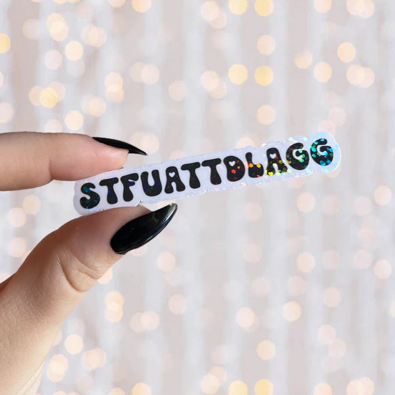 May include: A white sticker with the text "STFUATTBLAGG" in black, bubble letters with rainbow glitter. The sticker is being held by a hand with black nail polish.