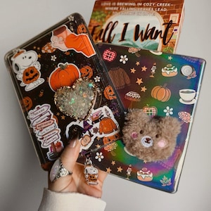 May include: Two tablet cases decorated with autumn-themed stickers including pumpkins, Snoopy, and cosy jumpers. One case features a heart-shaped glitter pop socket, while the other has a fuzzy brown bear attachment and a small Snoopy charm.