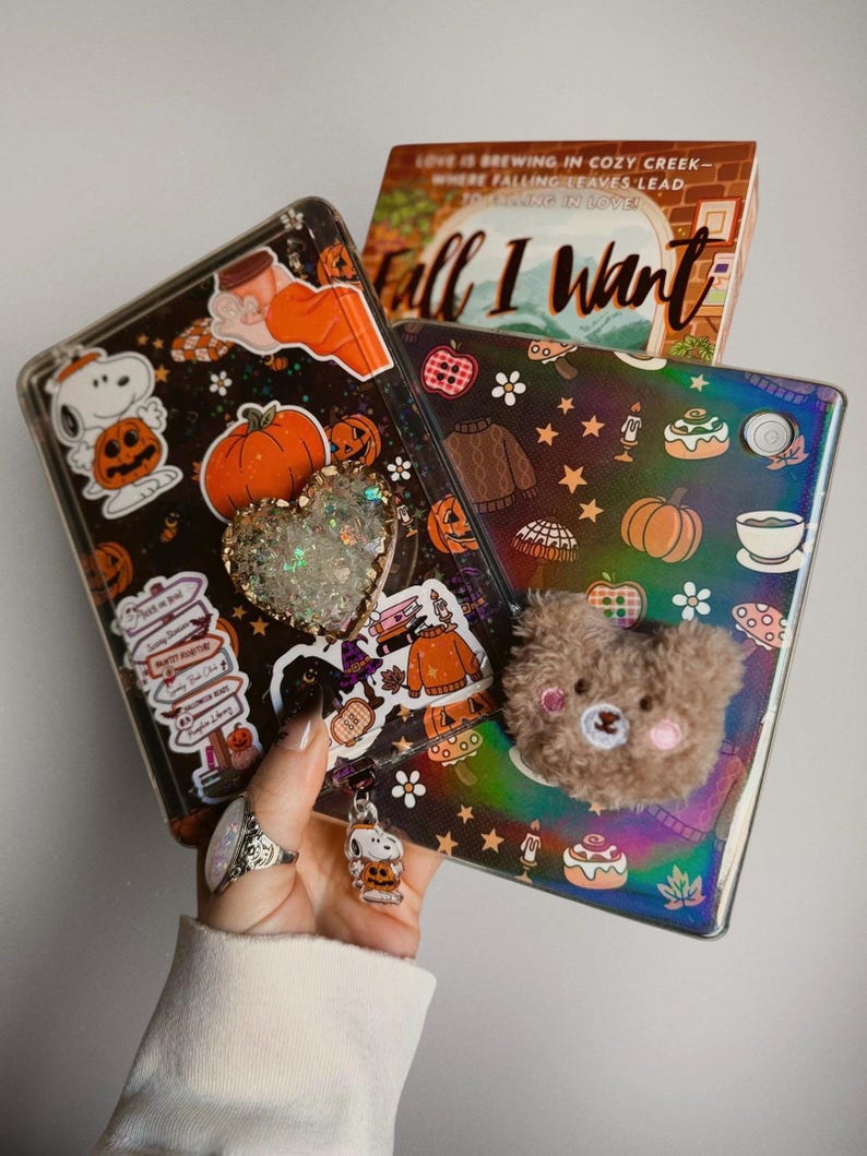 May include: Two tablet cases decorated with autumn-themed stickers including pumpkins, Snoopy, and cosy jumpers. One case features a heart-shaped glitter pop socket, while the other has a fuzzy brown bear attachment and a small Snoopy charm.