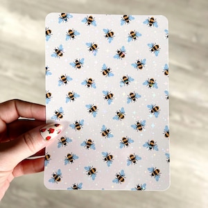 Puede incluir: Una hoja de papel blanca con un patrón repetitivo de abejas azules y amarillas sobre un fondo blanco con un sutil efecto de brillo.