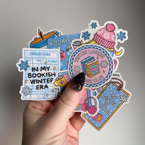 May include: A collection of winter-themed stickers featuring a pink and white beanie, books, snowflakes, and the text "Winter Lovers Book Club." Other stickers include a library card and the phrase "In My Bookish Winter Era."