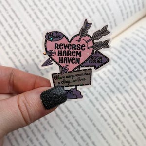 May include: A pink heart-shaped sign with the text "Reverse Harem Haven" and "Room for All". The sign is decorated with arrows and glitter. The text below the sign reads "Where every room has a king...or three."