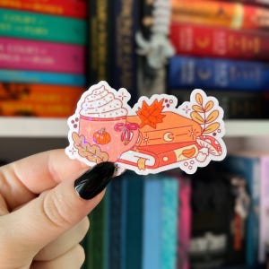 May include: A colorful sticker featuring a fall theme. It depicts a mug of whipped cream, a pumpkin, two books, a maple leaf, and a mushroom. The sticker has a holographic effect and is in shades of orange, pink, and white.