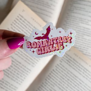 May include: A white sticker with a pink and white design that says "ROMantasy Girlie" with a silhouette of a bat in the background. The sticker has a holographic finish.