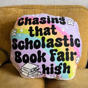 May include: A white pillow with a rainbow and the text "Chasing that Scholastic Book Fair high" in black. A stack of books is pictured below the text.