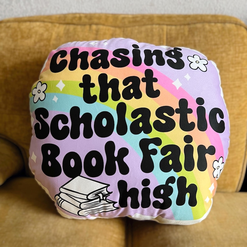 May include: A white pillow with a rainbow and the text "Chasing that Scholastic Book Fair high" in black. A stack of books is pictured below the text.