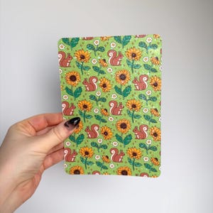 May include: A rectangular card with a green background featuring a repeating pattern of sunflowers, squirrels, and small white flowers. The sunflowers are yellow and orange, and the squirrels are brown. The card is held by a hand.