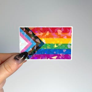 May include: A rectangular sticker featuring the Progress Pride flag design with horizontal stripes of red, orange, yellow, green, blue, and violet, alongside a chevron of black, brown, light blue, pink, and white, all with a holographic finish.