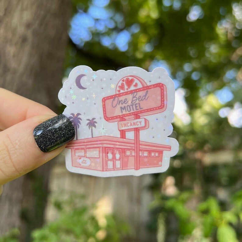 May include: A pink and white sticker with a retro motel sign that reads "One Bed Motel" with a vacancy sign. The sticker has a glittery finish and features a crescent moon and stars.