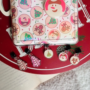 May include: A clear phone case featuring a repeating pattern of circular icons with Christmas trees, reindeer, and snowmen. Five small, circular and tree-shaped charms with matching holiday graphics are attached to the bottom of the case.